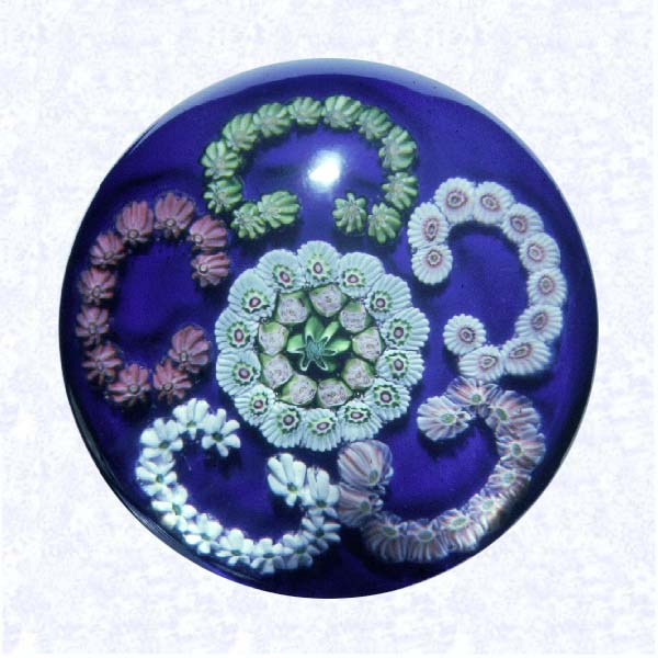 <B>&quotC" Garland Millefiori on Blue<BR>France<BR>Clichy, circa 1845-55</B><BR>Diameter: 7 cm (2 3/4 inches)<BR>(702416)<BR><BR>&quotC" scroll garland millefiori with five scrolls in green, pink, and white, encircling two concentric rings; inner ring comprised of nine pink Clichy roses around a green center cane; on a dark blue opaque ground